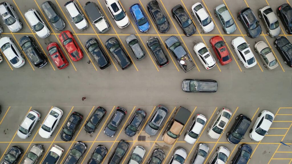 Overhead aerial view of a full parking lot with rows of cars in marked spaces and one vehicle moving along the open center aisle